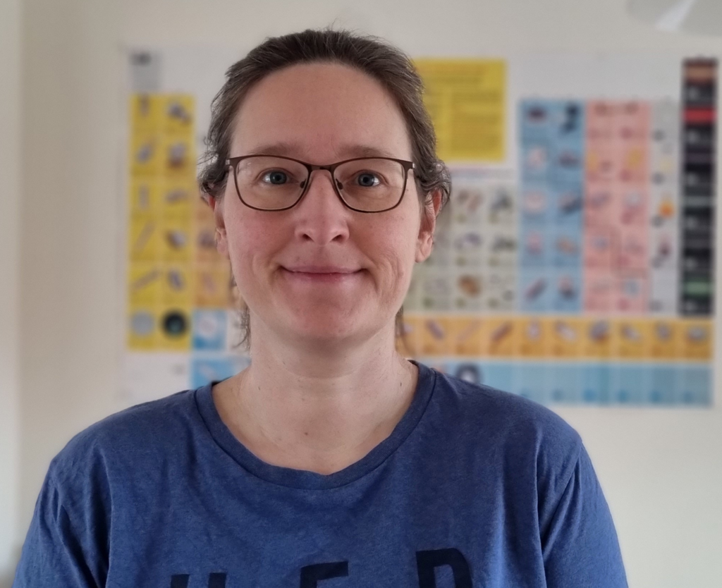 tutoring with angela: close up porrait of a woman smiling into the camera. in the blurred background there is a poster of the periodic table of the elements on the wall