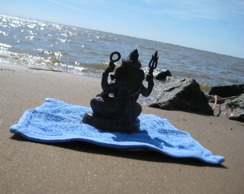 as long as: photo of a small Ganesh staue on a blue towel at the beach. the water in the background glitters with the sunlight