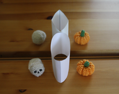 at the same time: photo of a crotcheted skull and a crotcheted pumpkin in front of a mirror. in between them is an object made from paper that looks like a cylinder from one direction and a cuboid from the other side