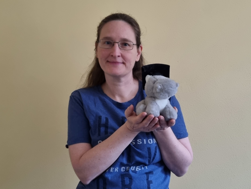 Woman holding a plush hippo in her hands. The hippo is wearing a doctorate hat. If you want to know about me, this is who I am as a teacher.