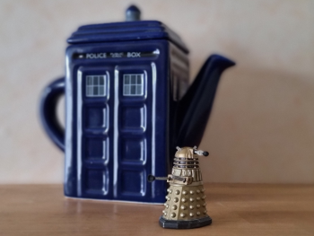 About me: a tea pot in the shape of a tardis with a small dalek model in front of it
